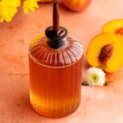 Peach simple syrup in a dispenser next to a flower and fresh peach.